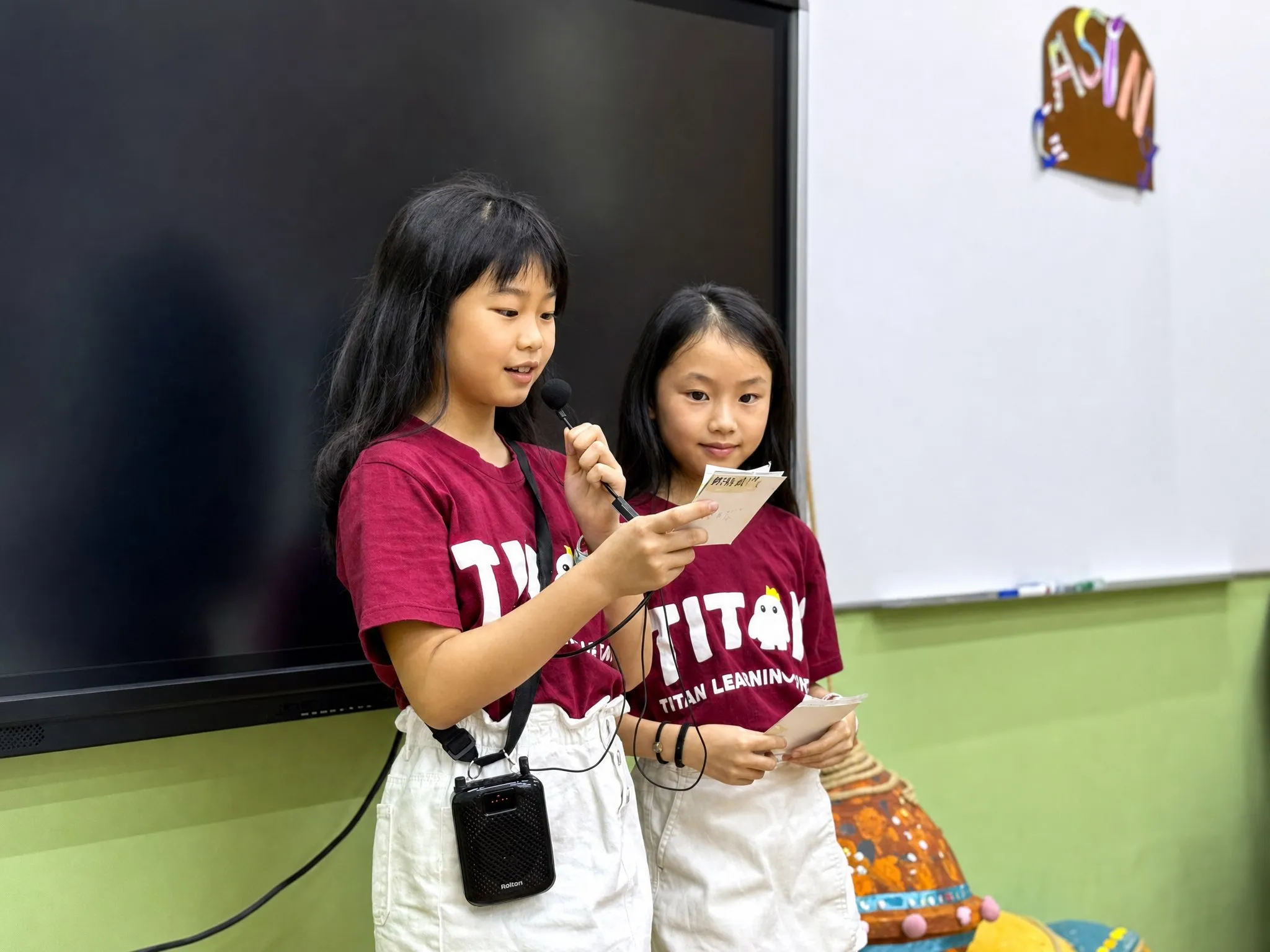 Two TITAN students presenting to their class, one speaking into a microphone while reading from a cue card