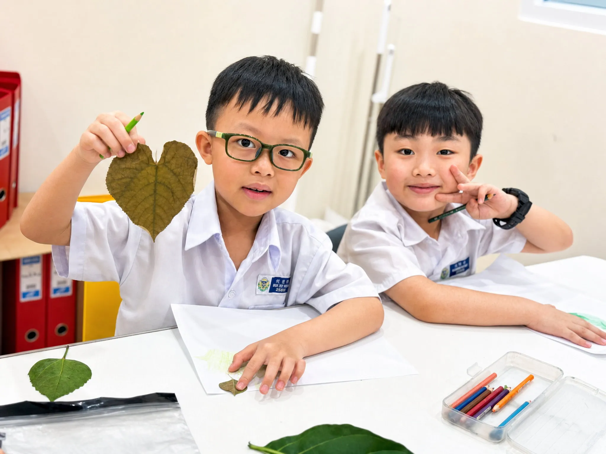 Two TITAN students at a table of leaves and coloured pencils, one holding up a dried leaf during a science activity